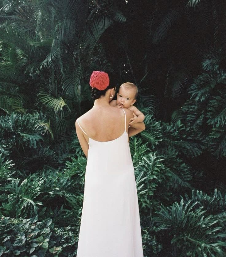 A woman in a white slip dress seen from behind, holding a baby against her shoulder in front of dense green foliage.